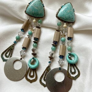 May include: A pair of statement earrings featuring turquoise triangular stones, wooden beads, and dangling silver-toned metal elements. The earrings have a mix of clear and turquoise beads, with a unique, bohemian design.