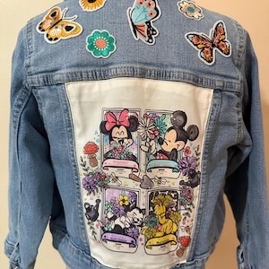 May include: A light blue denim jacket with a white patch on the back. The patch features four cartoon characters surrounded by flowers and other nature-themed designs. The jacket has several colorful embroidered patches on the front, including butterflies, flowers, and a sunflower.