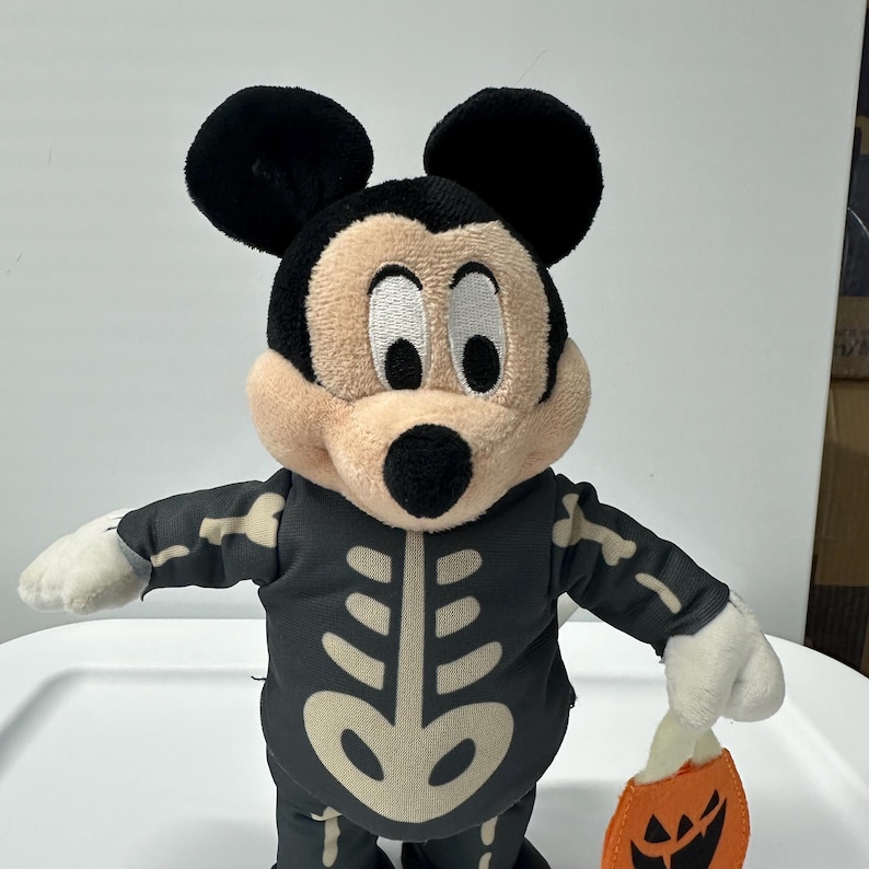 以下が含まれることがあります： ハロウィーンのスケルトンコスチュームを着たミッキーマウスのぬいぐるみ。黒い衣装には白いスケルトンデザインがあります。ミッキーはオレンジ色のカボチャ型のトリックオアトリートバッグを持っています。おもちゃは黒い耳、日焼けした顔、白い手袋をしています。