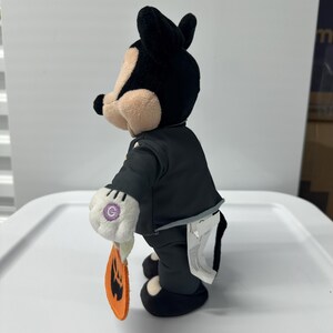 以下が含まれることがあります： 黒いスーツ、黒い靴、白い手袋を着たミッキーマウスのぬいぐるみ。このフィギュアは、ジャック・オ・ランタンの顔が付いたオレンジ色のカボチャ型のタグを持っています。黒い耳、黒い鼻、日焼けした頬をしています。