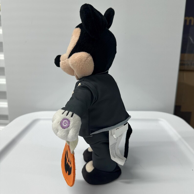 以下が含まれることがあります： 黒いスーツ、黒い靴、白い手袋を着たミッキーマウスのぬいぐるみ。このフィギュアは、ジャック・オ・ランタンの顔が付いたオレンジ色のカボチャ型のタグを持っています。黒い耳、黒い鼻、日焼けした頬をしています。