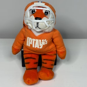 May include: Orange plush tiger wearing a white football helmet and orange jersey with the text "IPTAY" and a paw print. The tiger has black stripes on its legs and is posed upright.