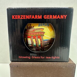 May include: A black box with the text "KERZENFARM GERMANY" in red. A circular window reveals a colorful illustration of a building with the word "BERLIN" above it. The text "Glowing Glass for tea-lights" is below the window.