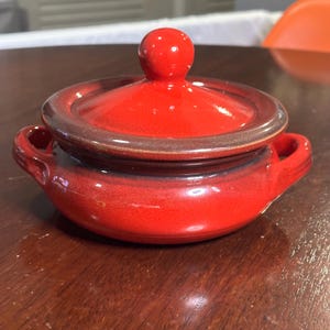 以下が含まれることがあります： 蓋付きの小さな赤い陶器のポット。ポットには2つのハンドルと濃い茶色の縁があります。蓋には小さな丸いノブが付いています。ポットは濃い木製の表面に置かれています。