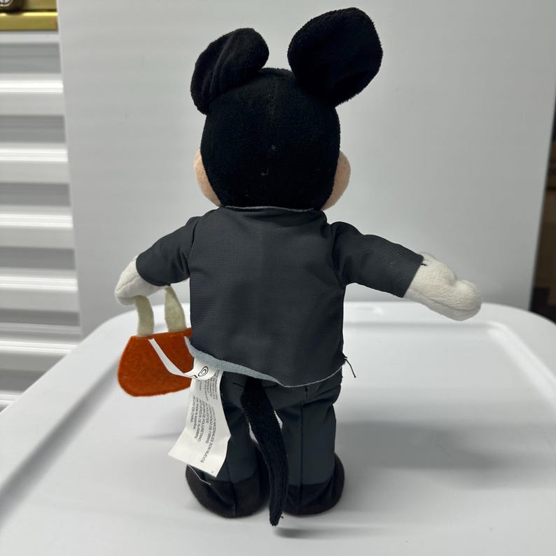 以下が含まれることがあります： 後ろから見たミッキーマウスのぬいぐるみ。黒いスーツを着て、小さなオレンジ色のハンドバッグを持っています。ぬいぐるみは黒い耳、黒い尻尾、白い手袋をしています。タグがぬいぐるみについています。