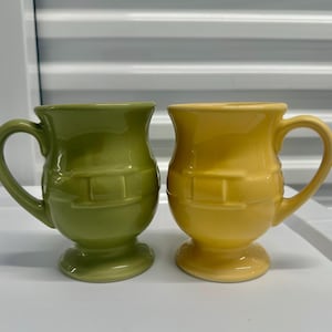 May include: Two ceramic mugs with a glossy finish, one in light green and one in yellow. Each mug features a rounded body, a pedestal base, and a handle on the side. The exterior of both mugs is decorated with a raised, horizontal band design.