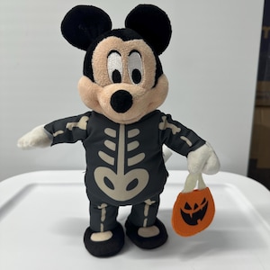 以下が含まれることがあります： 黒いスケルトンコスチュームを着たミッキーマウスのぬいぐるみ。ぬいぐるみはベージュの顔、黒い耳をしており、ジャック・オー・ランタンの顔が付いたオレンジ色のトリックオアトリートバッグを持っています。ハロウィーンをテーマにしたコレクターズアイテム。