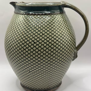 May include: A green ceramic pitcher with a textured diamond pattern. The pitcher has a wide base and a rounded body. The handle is attached to the side of the pitcher.