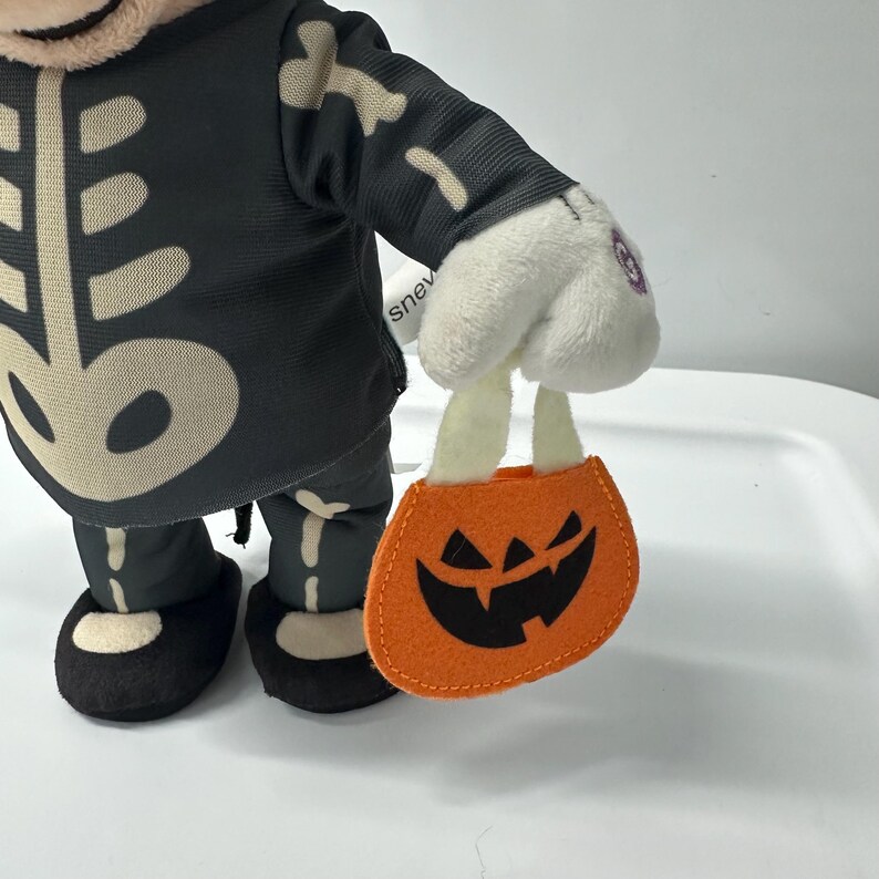 以下が含まれることがあります： 骨色のアクセントが付いた黒いスケルトンコスチュームを着たぬいぐるみ。おもちゃは、ジャック・オ・ランタンの顔が付いたオレンジ色のフェルトカボチャ型のトリックオアトリートバッグを持っています。おもちゃの手袋は白です。