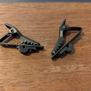 Puede incluir: Dos pinzas de corbata de metal negro se muestran sobre una superficie de madera. Cada pinza tiene un diseño articulado con un borde dentado para sujetar la tela. Las pinzas están diseñadas para asegurar una corbata a una camisa.