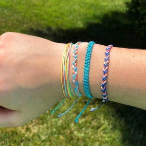 May include: A set of five colorful friendship bracelets, featuring a mix of blue, yellow, and pink woven threads. The bracelets are tied together and worn on a wrist.