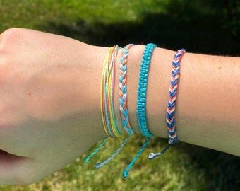 May include: A set of five colorful friendship bracelets, featuring a mix of blue, yellow, and pink woven threads. The bracelets are tied together and worn on a wrist.