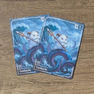 May include: Two rectangular game cards featuring a white, furry creature with small horns holding a golden trident. The creature stands atop a large, dark purple octopus tentacle amidst turbulent blue ocean waves. The word Tentacle is printed at the bottom.