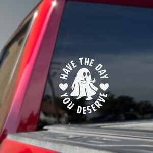 May include: White vinyl sticker with a ghost giving a thumbs up and the text "Have the day you deserve" in a circle with hearts on either side of the ghost.