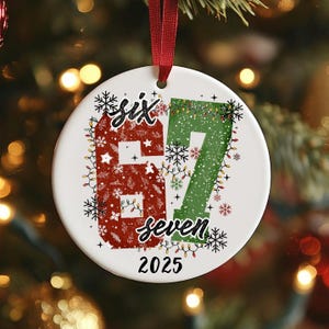 May include: A white ceramic Christmas ornament with a red ribbon. The ornament features the words "six" and "seven" with the numbers 6 and 7. The number 6 is red with a snowflake pattern, and the number 7 is green. The year 2025 is at the bottom.