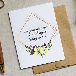 May include: A white card with a gold geometric frame and floral design. The text inside the frame reads "congratulations on no longer living in sin".