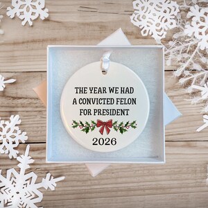 May include: A round white ceramic ornament featuring the black text THE YEAR WE HAD A CONVICTED FELON FOR PRESIDENT, a red bow, green holly leaves, and the year 2026. The ornament includes a white ribbon loop and is presented in a white gift box.