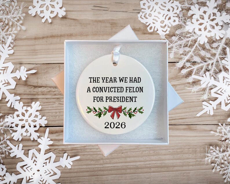 May include: A round white ceramic ornament featuring the black text THE YEAR WE HAD A CONVICTED FELON FOR PRESIDENT, a red bow, green holly leaves, and the year 2026. The ornament includes a white ribbon loop and is presented in a white gift box.