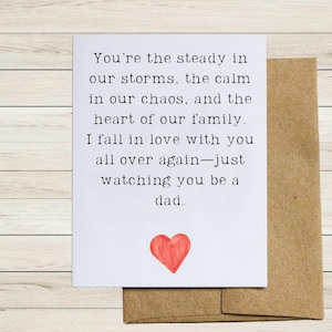 May include: A white greeting card with black text and a red heart illustration. The card is placed on top of a brown envelope. The text reads: "You're the steady in our storms, the calm in our chaos, and the heart of our family. I fall in love with you all over again-just watching you be a dad."