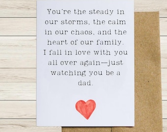 May include: A white greeting card with black text and a red heart illustration. The card is placed on top of a brown envelope. The text reads: "You're the steady in our storms, the calm in our chaos, and the heart of our family. I fall in love with you all over again-just watching you be a dad."