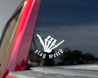 May include: White vinyl decal featuring a skeleton hand in the shaka sign, with the words "stay weird" in a gothic font. The decal is applied to the rear window of a red truck.
