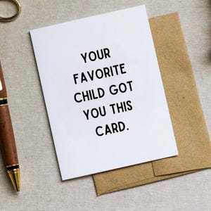 May include: A white greeting card with the text "YOUR FAVORITE CHILD GOT YOU THIS CARD" in black, placed on a brown envelope. A gold pen and a gold ring are visible in the background.