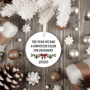 May include: A round white ceramic Christmas ornament hanging from a white ribbon. The ornament features black text that reads THE YEAR WE HAD A CONVICTED FELON FOR PRESIDENT, a red bow graphic with green holly leaves, and the year 2026 printed below.