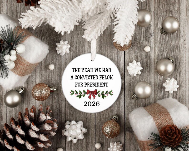 May include: A round white ceramic Christmas ornament hanging from a white ribbon. The ornament features black text that reads THE YEAR WE HAD A CONVICTED FELON FOR PRESIDENT, a red bow graphic with green holly leaves, and the year 2026 printed below.