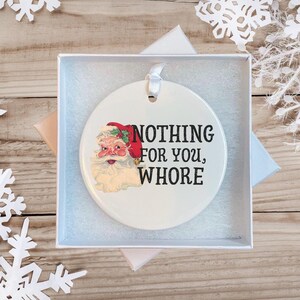 May include: White ceramic Christmas ornament with a cartoon image of Santa Claus and the text "NOTHING FOR YOU, WHORE".