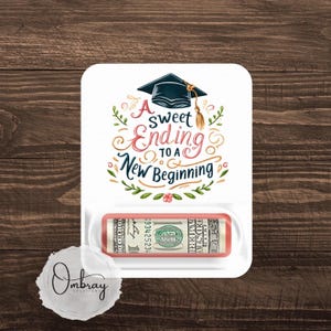 May include: A white card with a graduation cap and floral design. The text reads "A sweet ending to a new beginning". A dollar bill is visible in a slot on the card.