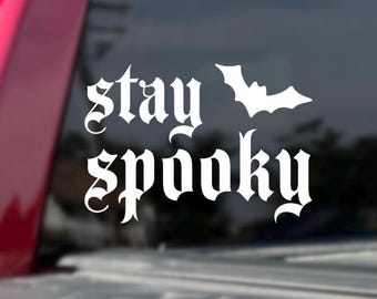 May include: White vinyl decal with the words "stay spooky" in a gothic font, accompanied by a bat silhouette. The decal is likely for a car window or similar surface, promoting a Halloween or spooky theme.