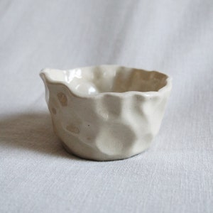 Puede incluir: Un pequeño cuenco de cerámica blanca con una superficie texturizada y ondulada. El cuenco es ligeramente irregular en forma y tiene un fondo redondeado.