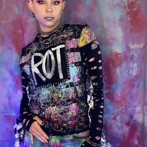 May include: A long-sleeve black top featuring the word ROT in white letters, layered over a colorful abstract print with distressed cutouts on the sleeves. The outfit includes tie-dye style pants with various graphic symbols and stitched details.