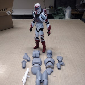 Puede incluir: Una figura de acción blanca y roja de un clon de Star Wars con un casco ensangrentado. La figura está desmontada y colocada sobre una superficie de madera. Las partes de la figura son grises y blancas.