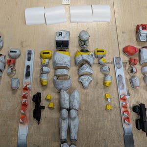 May include: A disassembled action figure, with various body parts including a helmet, torso, limbs, and accessories. The figure is mainly grey with yellow and red accents. The parts are arranged on a wooden surface.