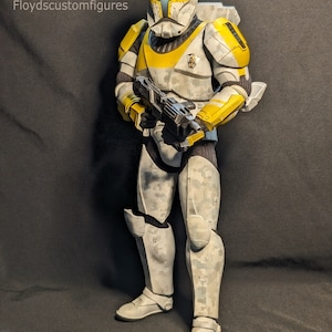 May include: A custom-painted Star Wars clone trooper action figure with a white and grey camouflage pattern, yellow armour accents, and a black blaster rifle. The figure is standing on a grey background. FCF Floyd's custom figures.