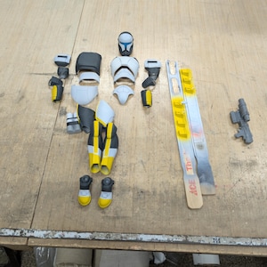 Op de afbeelding: Een gedemonteerd actiefiguur van een Star Wars kloonstrijder. De figuur is wit, zwart en geel geverfd. De figuur ligt op een houten oppervlak.