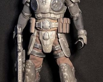 May include: A detailed action figure of a soldier wearing metallic gray tactical armor with a skull emblem on the chest plate. The figure wears a dark cap, brown trousers, and carries a long rifle in one hand, featuring weathered textures.