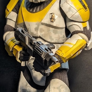 Peut inclure: Une figurine de collection représentant un soldat portant une armure blanche et jaune avec un motif camouflage numérique. La figurine possède un casque à visière bleue, une plaque de poitrine avec un emblème noir et or, et tient un fusil blaster noir.