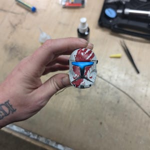 Puede incluir: Un casco blanco con detalles de pintura roja y azul. El casco tiene una visera azul y una huella de mano roja en la parte delantera.