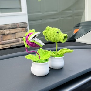 May include: Two crocheted plant figurines in white pots. The plants are green and have a cartoonish appearance. One plant has a large, open mouth with white teeth and a purple tongue. The other plant has a round head with black eyes and a green stem.