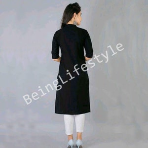 May include: A black long-sleeved tunic with a collar and a button fastening. The tunic is knee-length and has a relaxed fit. The model is wearing white trousers and blue shoes.