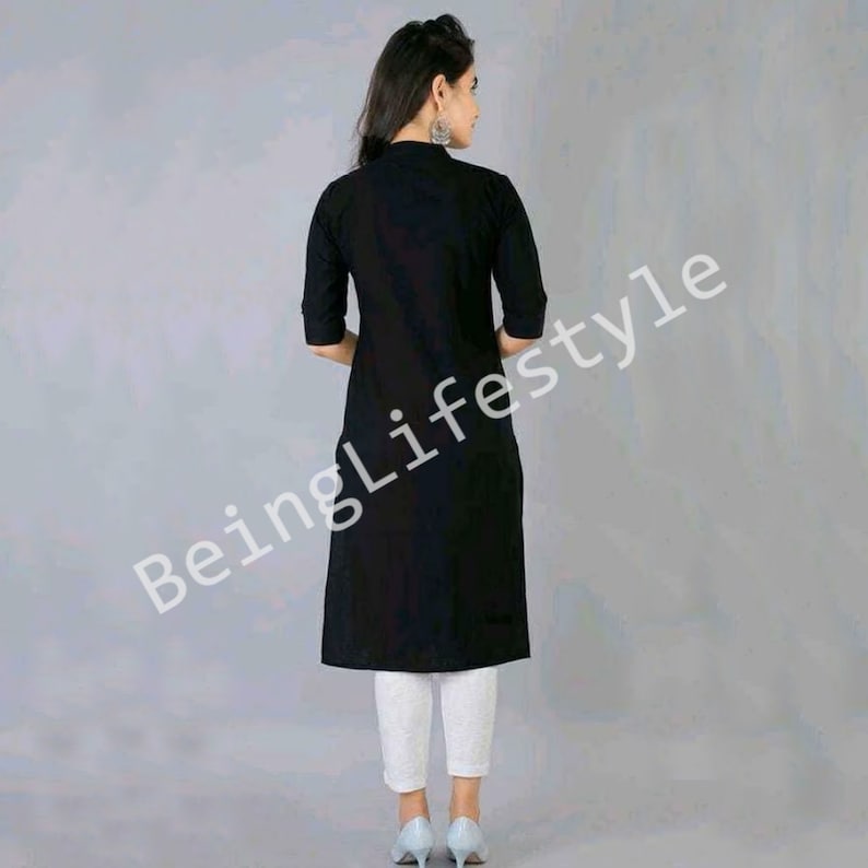 May include: A black long-sleeved tunic with a collar and a button fastening. The tunic is knee-length and has a relaxed fit. The model is wearing white trousers and blue shoes.