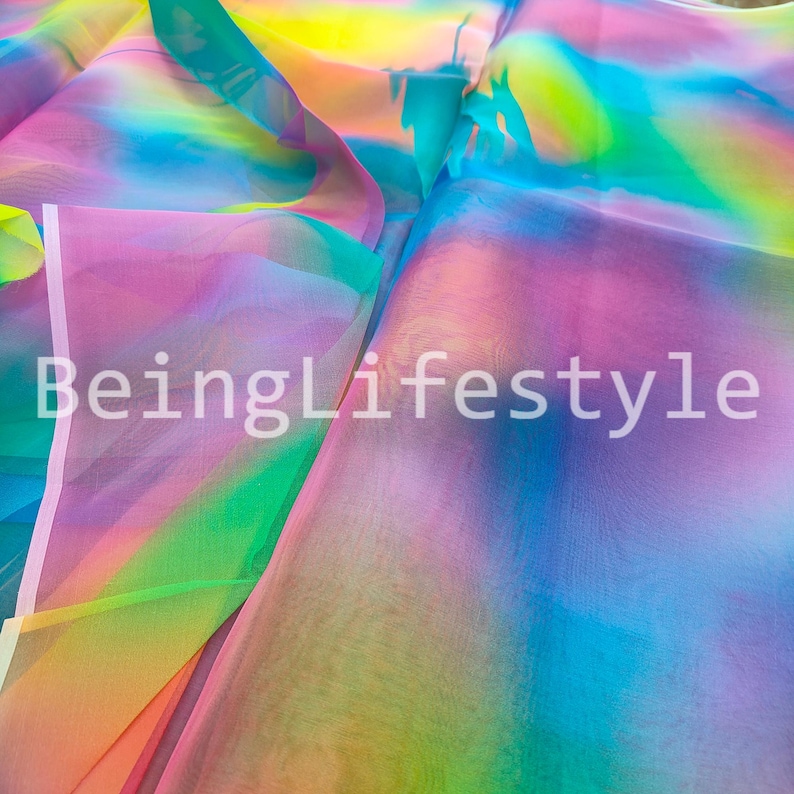 May include: A close-up of a sheer fabric with a rainbow gradient of colors, including pink, yellow, green, and blue. The fabric is folded and layered, creating depth and texture. The text "BeingLifestyle" is visible in white.
