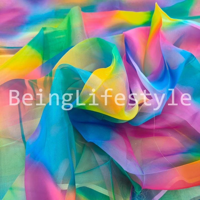 May include: A vibrant, flowing fabric in a rainbow of colors, including blue, green, yellow, pink, and orange. The text "BeingLifestyle" is visible on the fabric. The material appears to be lightweight and translucent, with soft folds and a silky texture.