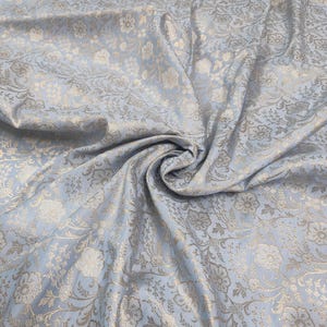 May include: A light blue fabric with a silver floral pattern. The fabric is woven with a subtle texture and has a soft, drapey feel.