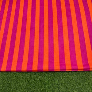 Könnte beinhalten: Ein lebendiger Stoff mit vertikalen Streifen in abwechselnden Orange- und Magentatönen. Der Stoff liegt auf einem Bett aus grünem Kunstrasen und erzeugt einen auffälligen Kontrast.