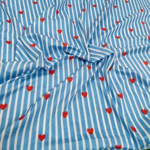 Puede incluir: Tela a rayas azules y blancas con un patrón de corazones rojos. La tela está recogida y doblada, mostrando la textura y el diseño. Los corazones están espaciados uniformemente sobre el material.