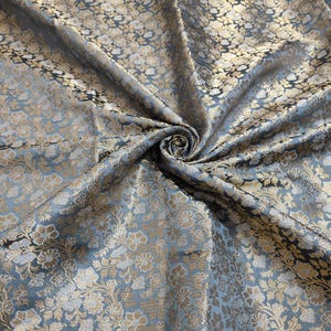 May include: A close-up of a blue and gold brocade fabric with a floral pattern. The fabric is folded and draped, showing the texture and detail of the design.
