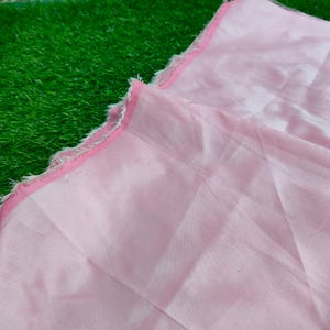 Pode incluir: Um tecido rosa claro com uma aparência delicada e translúcida. As bordas são adornadas com uma borda rosa contrastante e uma franja de fios brancos. O tecido está dobrado, revelando sua textura e brilho. O fundo é verde vibrante.
